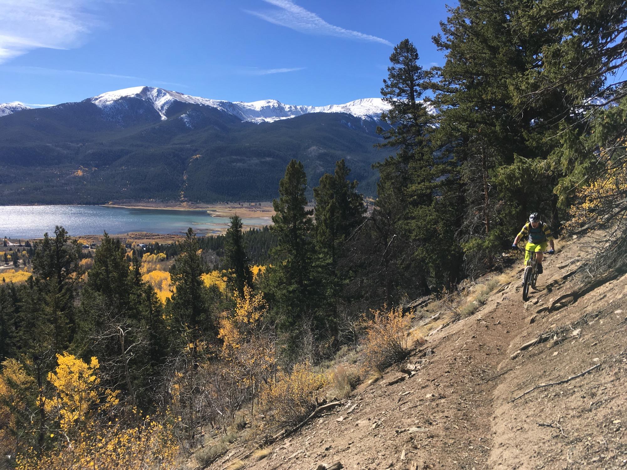 Mountain biker navigating a rocky trail surrounded by pine trees, with a stunning view of a lake and snow-capped mountains in the background on a clear autumn day. Colorado Trail: Twin Lakes / Hwy 82 to Half Moon Rd / Mount Massive Wilderness mountain bike trail.