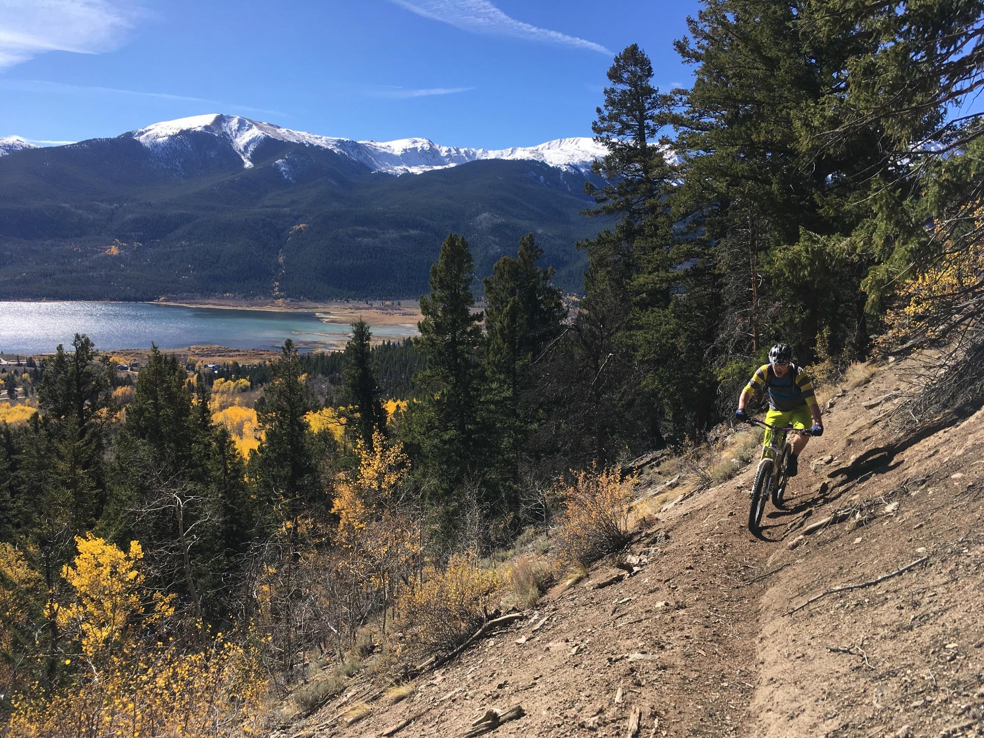 A mountain biker riding along a dirt trail surrounded by tall pine trees, with a view of a lake and mountains in the background. The scene is set in autumn, showcasing vibrant yellow foliage and snow-capped peaks under a clear blue sky. Colorado Trail: Twin Lakes / Hwy 82 to Half Moon Rd / Mount Massive Wilderness mountain bike trail.