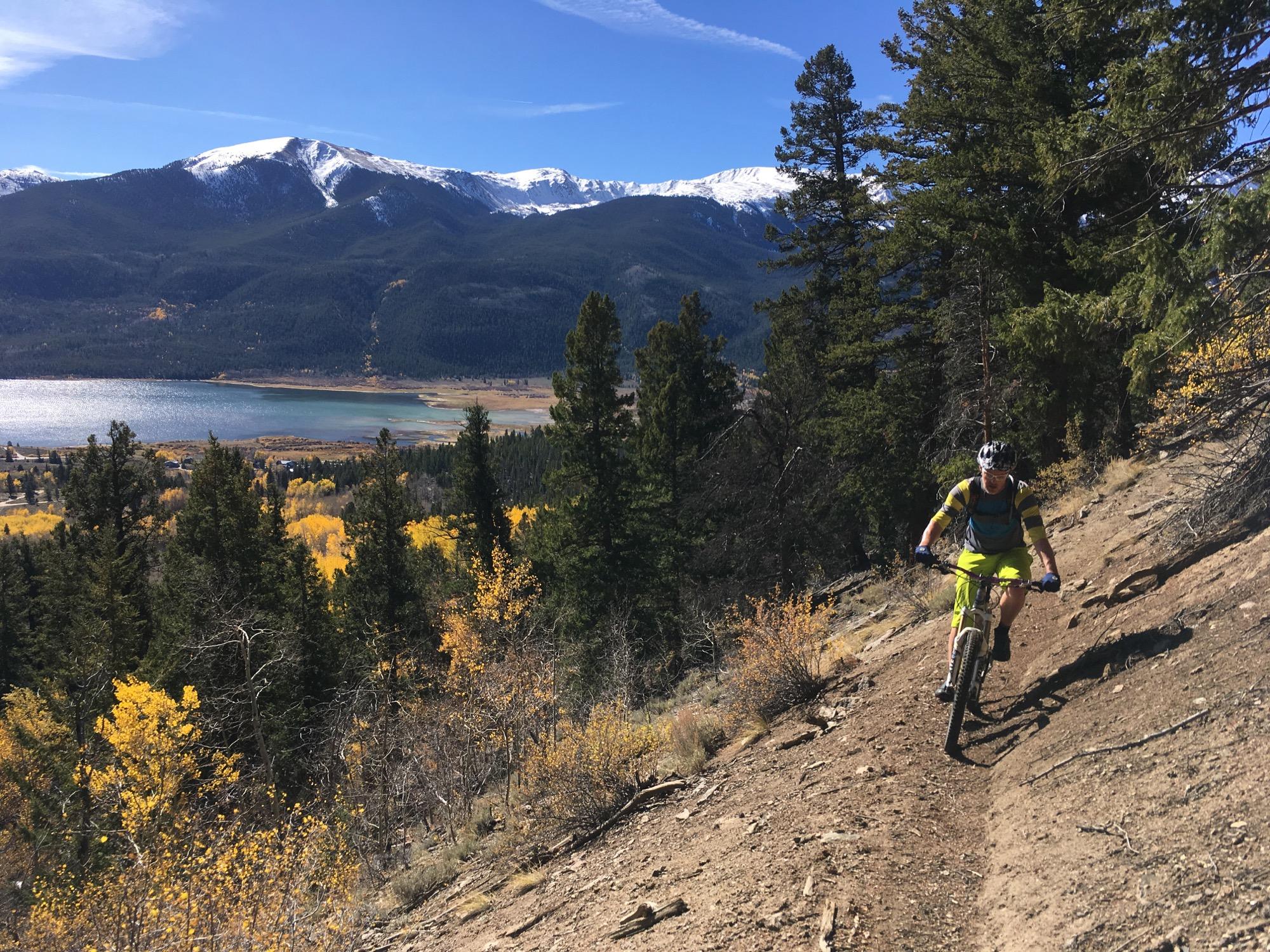 A mountain biker navigates a rugged trail surrounded by towering pine trees, with a panoramic view of a lake and snow-capped mountains in the background. The scene showcases vibrant autumn foliage and clear blue skies. Colorado Trail: Twin Lakes / Hwy 82 to Half Moon Rd / Mount Massive Wilderness mountain bike trail.