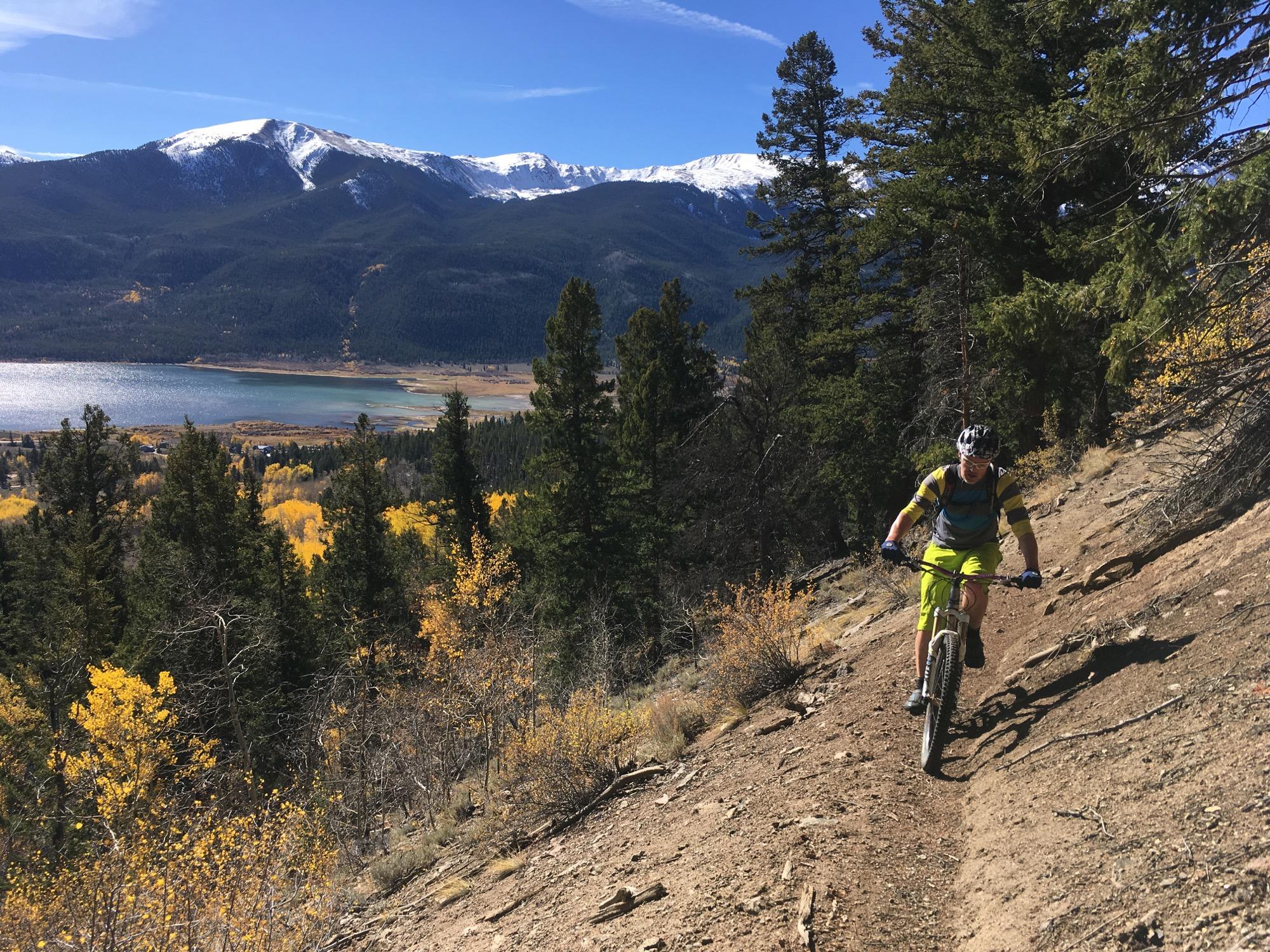 A mountain biker navigating a narrow dirt trail amidst a backdrop of majestic mountains and a serene lake. The trail is lined with tall evergreen trees and vibrant autumn foliage, showcasing shades of yellow against the rocky terrain. The sky is clear with a few wispy clouds, and snow-capped peaks are visible in the distance. Colorado Trail: Twin Lakes / Hwy 82 to Half Moon Rd / Mount Massive Wilderness mountain bike trail.