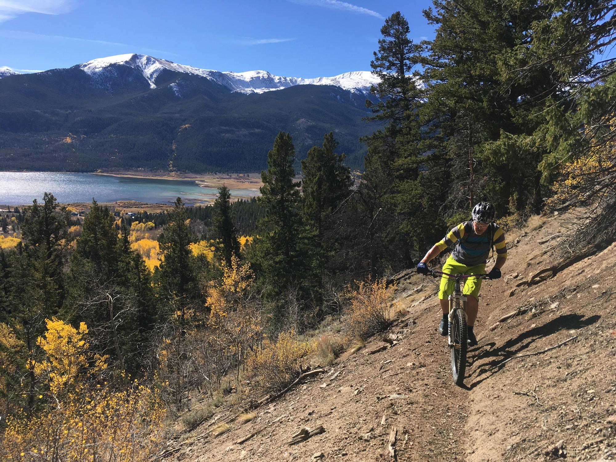 A mountain biker navigating a narrow trail in a forested area, surrounded by tall trees and vibrant autumn foliage. In the background, a serene lake and snow-capped mountains create a picturesque landscape under a clear blue sky. Colorado Trail: Twin Lakes / Hwy 82 to Half Moon Rd / Mount Massive Wilderness mountain bike trail.