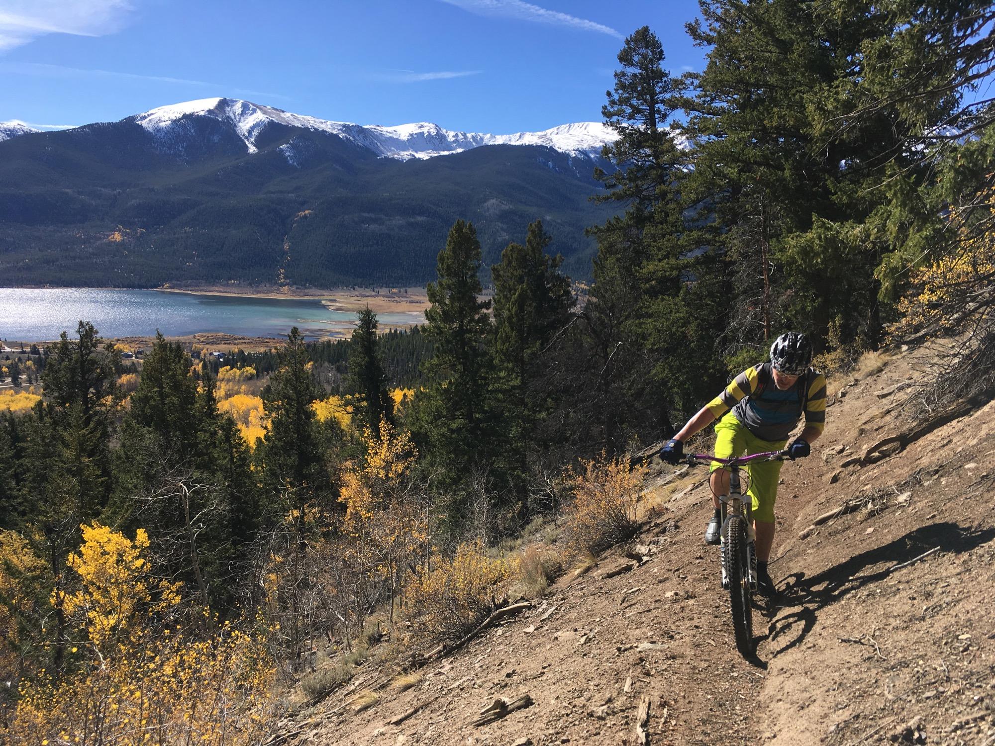 A mountain biker navigating a dirt trail on a hillside, surrounded by tall pine trees and colorful autumn foliage, with a clear blue sky and snow-capped mountains in the background. A lake can be seen below, reflecting the vibrant fall colors of the landscape. Colorado Trail: Twin Lakes / Hwy 82 to Half Moon Rd / Mount Massive Wilderness mountain bike trail.