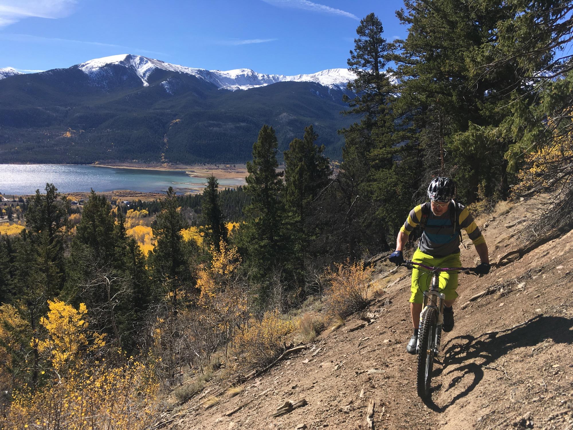 A mountain biker navigating a rocky trail in a forested area with autumn foliage. In the background, a lake and majestic snow-capped mountains under a clear blue sky. Colorado Trail: Twin Lakes / Hwy 82 to Half Moon Rd / Mount Massive Wilderness mountain bike trail.