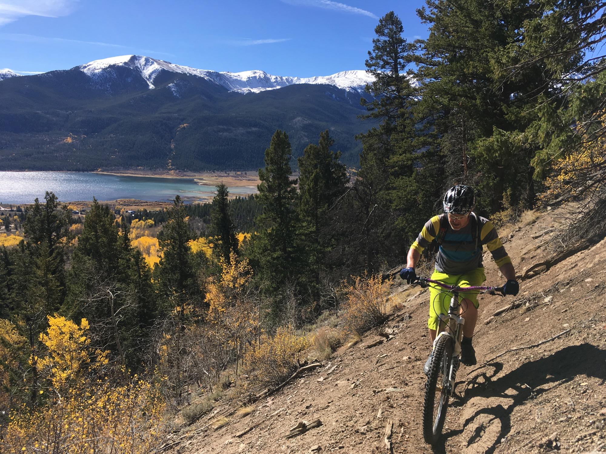 A mountain biker riding up a steep, rocky trail surrounded by pine trees, with a scenic view of mountains and a lake in the background. The foreground features golden autumn foliage, and the sky is clear with a few clouds. Colorado Trail: Twin Lakes / Hwy 82 to Half Moon Rd / Mount Massive Wilderness mountain bike trail.