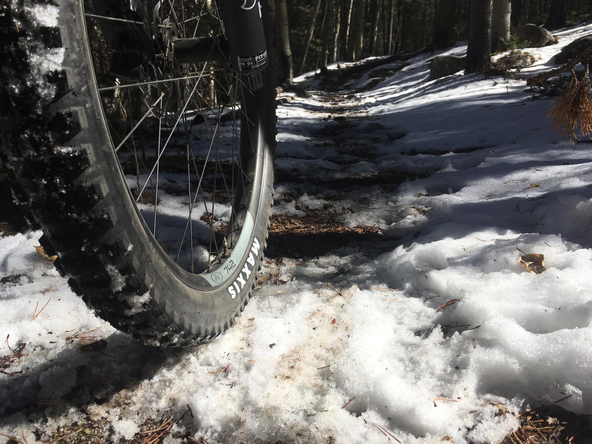 A close-up view of a mountain bike tire on a snow-covered trail in a forest. The surrounding area shows patches of snow and pine needles, with trees visible in the background. The sunlight is shining through the trees, creating a serene winter atmosphere. Colorado Trail: Mt. Shavano thd to Chalk Creek thd mountain bike trail.