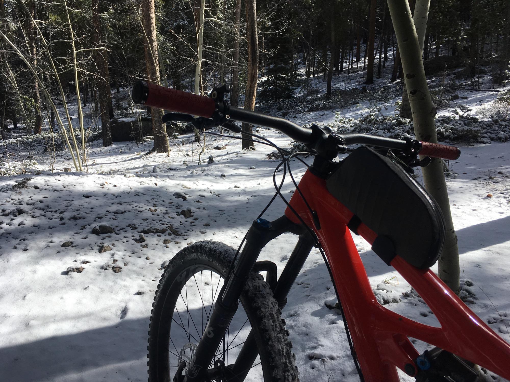A close-up view of a mountain bike in a snowy forest. The bike's frame is red, and the handlebars feature red grips. The background shows a landscape covered in a thin layer of snow, with trees and scattered rocks partially visible. Sunlight filters through the trees, creating a serene winter scene. Rainbow Trail: Silver Creek to Hwy 285 mountain bike trail.