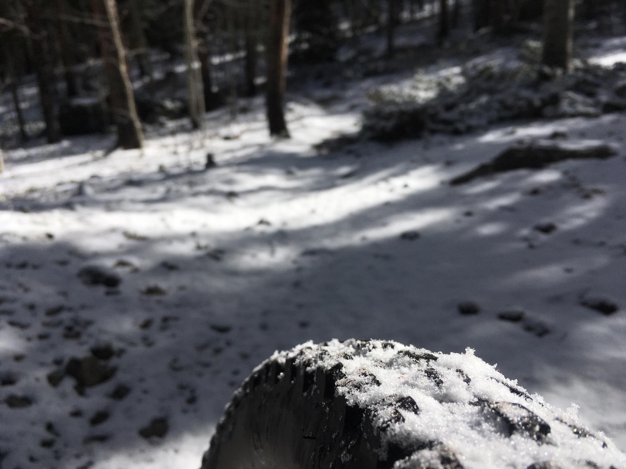 A close-up view of a tire partially covered in snow, with a snowy forest path in the background. Shadows from trees create a dappled light effect on the snow-covered ground. Rainbow Trail: Silver Creek to Hwy 285 mountain bike trail.