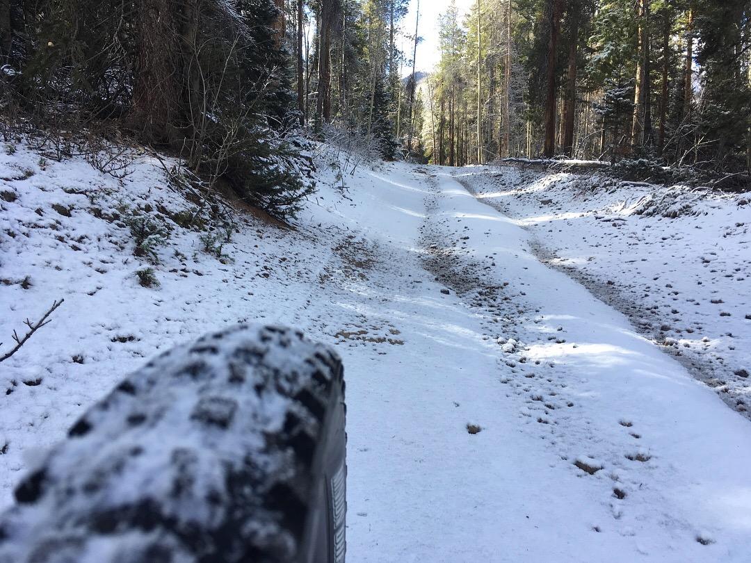 A snow-covered dirt road meanders through a forest, flanked by tall trees. In the foreground, a tire with a visible tread pattern partially obscures the view, indicating recent travel. The sun shines through the trees, creating a serene atmosphere in this winter landscape. Rainbow Trail: Silver Creek to Hwy 285 mountain bike trail.