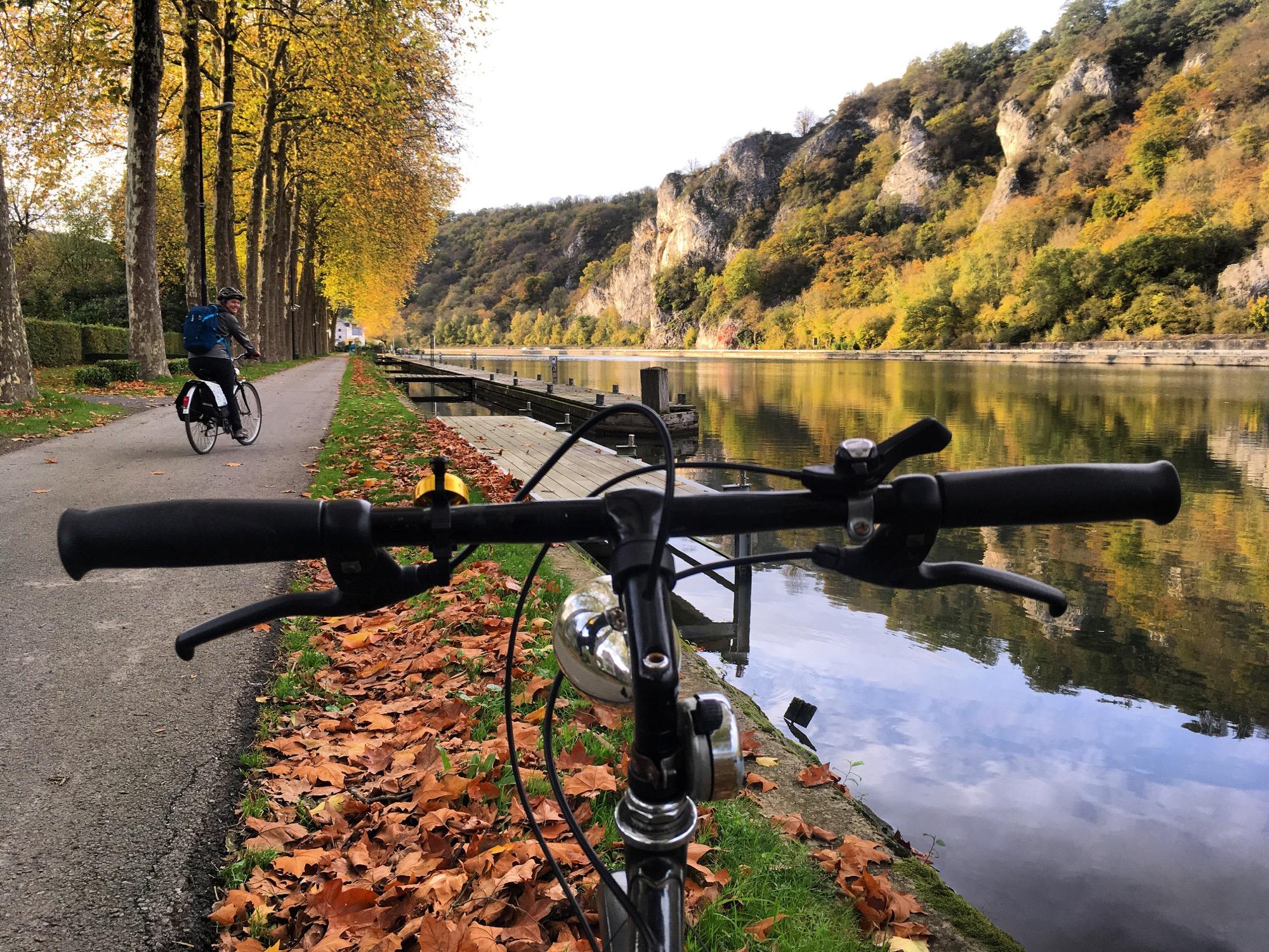 A view from the handlebars of a bicycle, with colorful autumn leaves on the ground. In the background, a pathway lined with trees runs alongside a calm river, reflecting the surrounding scenery. Another cyclist rides on the path, enjoying the peaceful outdoor setting. GR126 Meuse Section mountain bike trail.