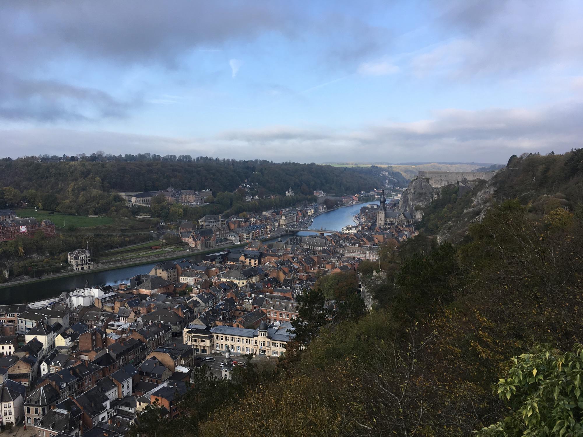 A panoramic view of a riverside town nestled between lush green hills. The scene features a winding river cutting through the landscape, with charming buildings in various architectural styles lining the banks. In the background, there are rolling hills and a rocky outcrop, partially shrouded in clouds, contributing to a serene and picturesque atmosphere. GR129S Dinant - Arlon mountain bike trail.