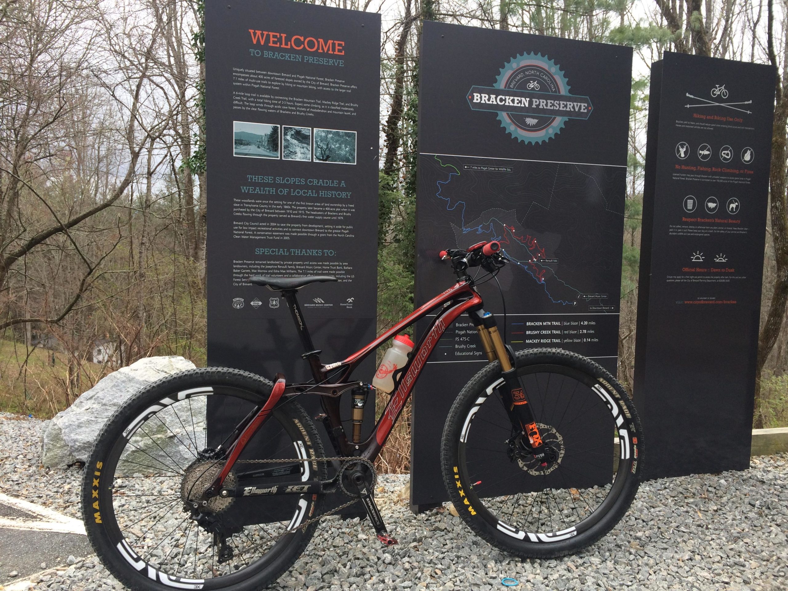 A mountain bike parked beside an informational sign at Bracken Preserve, detailing local history and trail options. The background features trees and gravel paths, with the sign showcasing a map and guidelines for visitors. Bracken Preserve mountain bike trail.