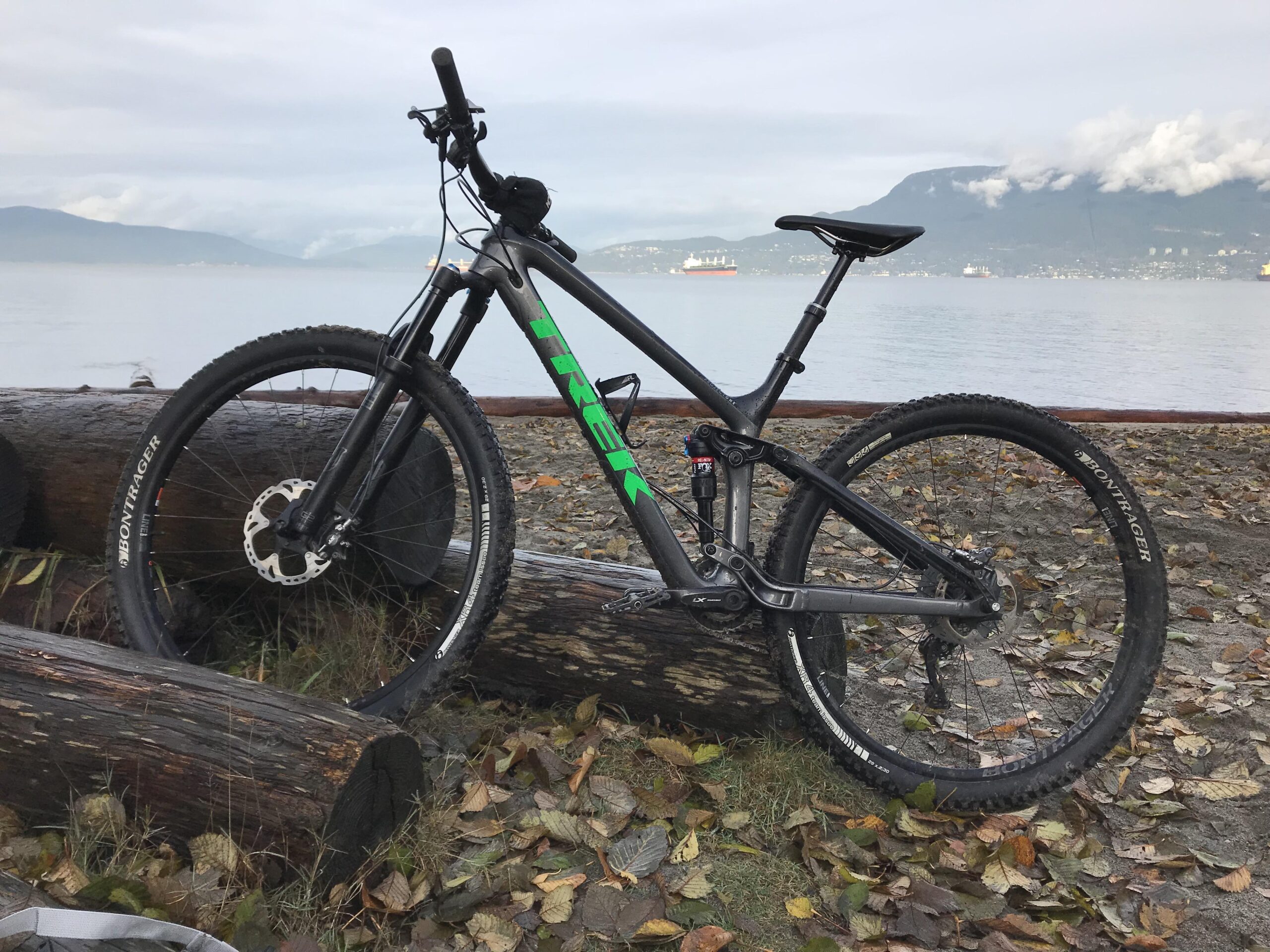 Trek Fuel EX 9.8: A mountain bike leaning against a log on a sandy shore, with calm water and distant mountains in the background. The bike features a black frame with green highlights and knobby tires, surrounded by fallen leaves.