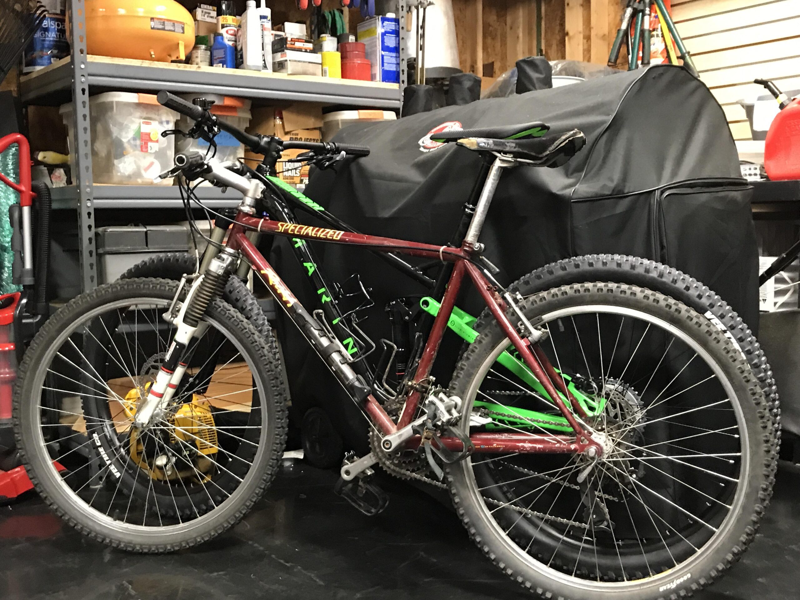 Specialized Rockhopper: A red and black mountain bike with green accents is leaning against a black covered object in a garage. Behind the bike, there are shelves filled with various tools and supplies, along with a vacuum cleaner and other items. The garage floor is dark, and the setting appears cluttered but organized.