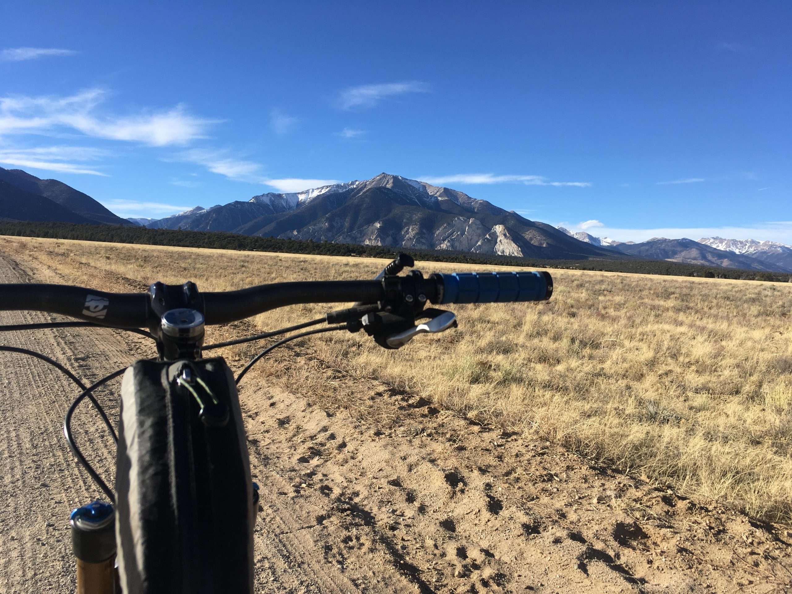 A view from the handlebars of a mountain bike, showing a dirt road leading towards mountains in the background. The sky is clear with a few clouds, and snow-capped peaks can be seen on the horizon. The foreground features dry grass and a rugged landscape typical of mountainous terrain. Browns Creek Road / #272 mountain bike trail.