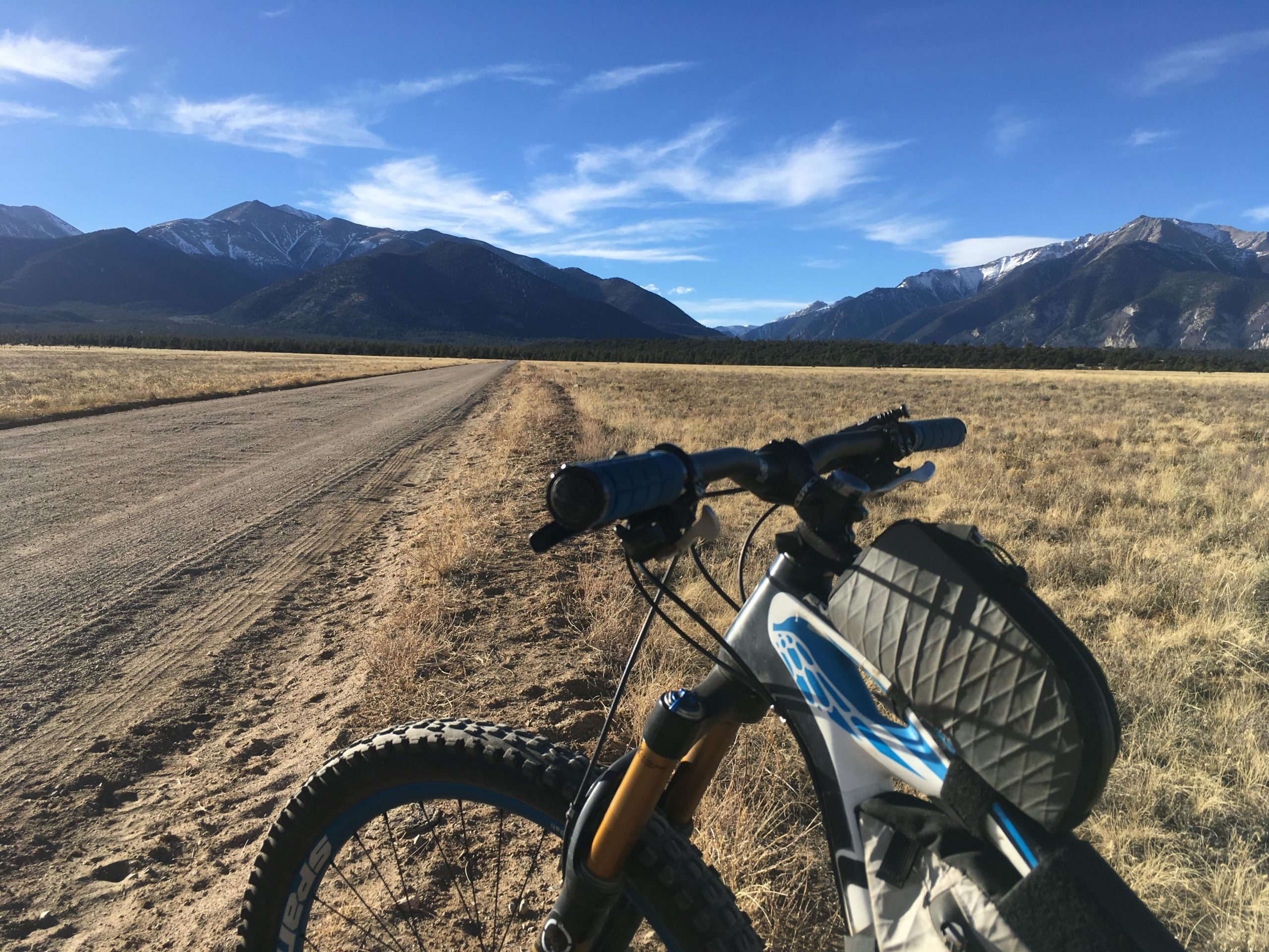 A mountain bike leaning on a dirt road with a scenic view of snow-capped mountains and a clear blue sky in the background, surrounded by open fields. Browns Creek Road / #272 mountain bike trail.