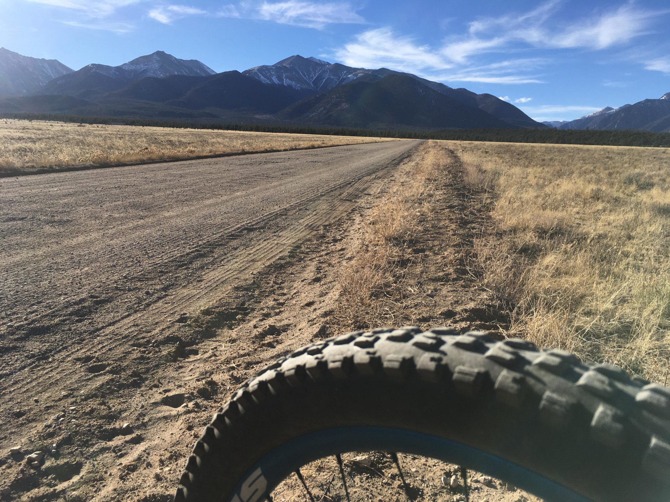 A close-up view of a bicycle tire resting on a dirt road, with a wide expanse of grassy land and distant snow-capped mountains under a blue sky. The scene captures the serene beauty of nature, ideal for outdoor activities such as biking or hiking. Browns Creek Road / #272 mountain bike trail.