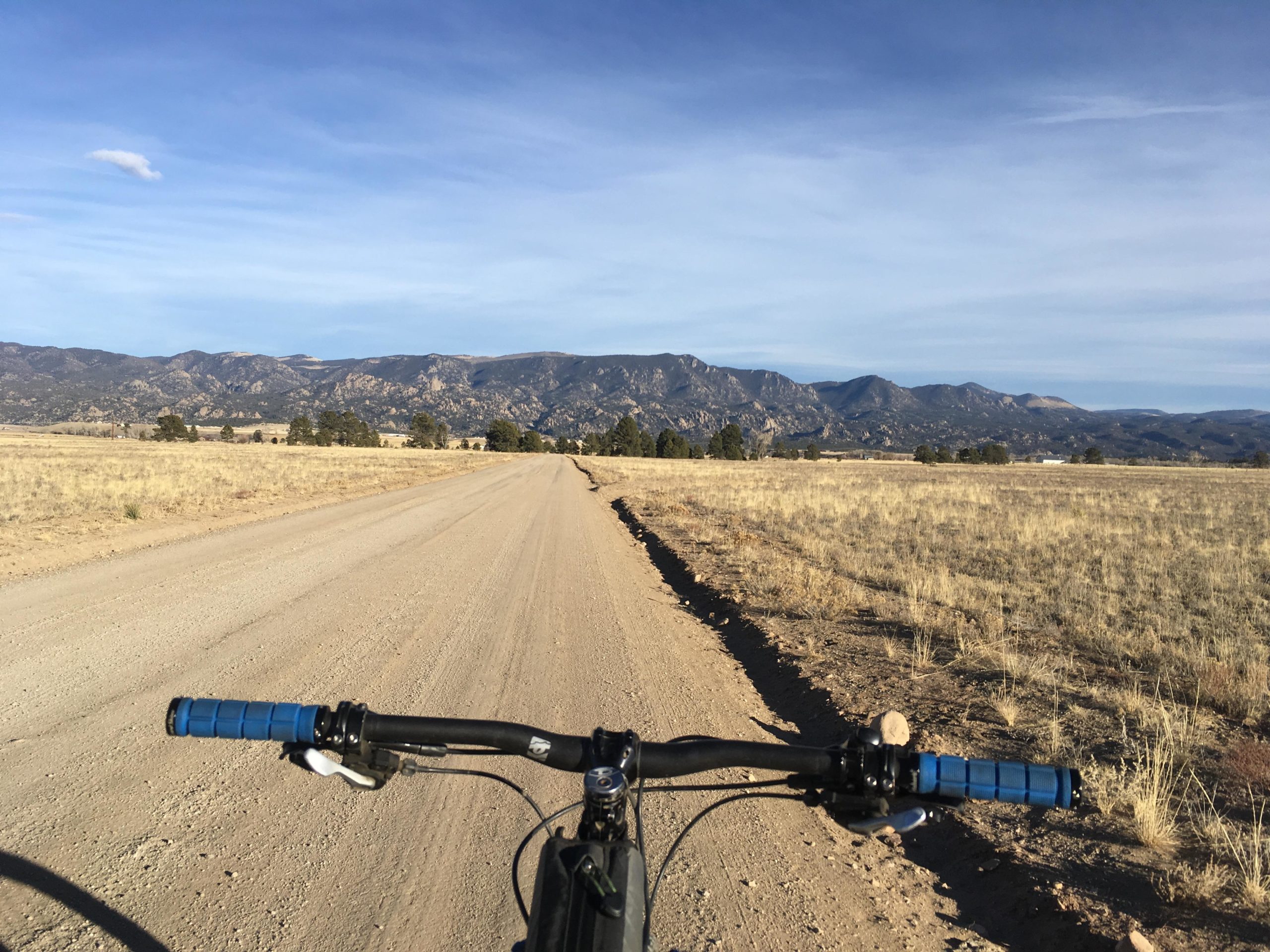 A view from the handlebars of a mountain bike on a dirt road, with expansive fields and a mountain range in the background under a blue sky. Browns Creek Road / #272 mountain bike trail.