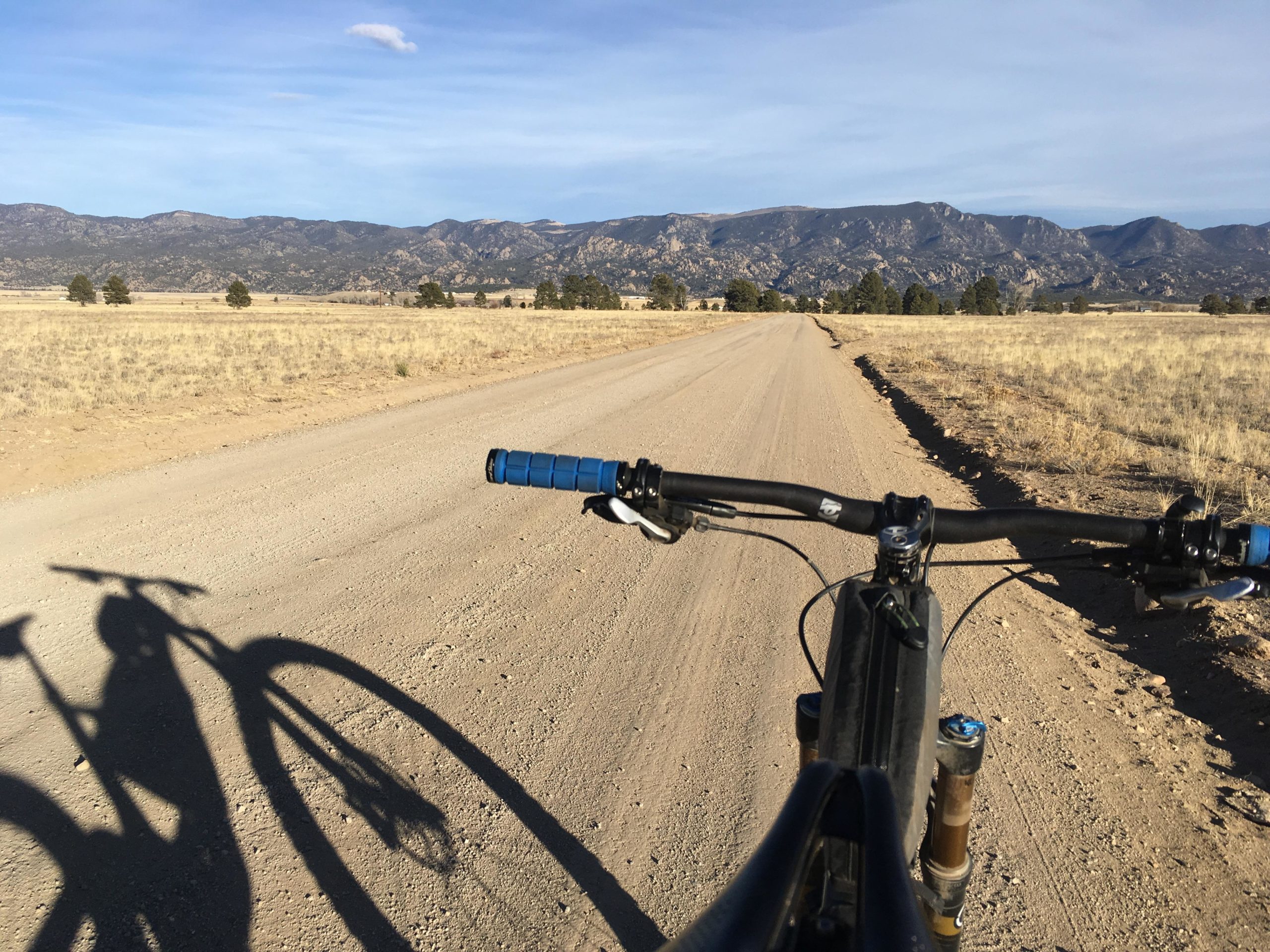 A view from the handlebars of a mountain bike on a dirt road, with a vast landscape of dry grass and sparse trees in the foreground. In the background, the rolling hills and mountains are visible under a blue sky, with a few clouds scattered above. The shadow of the bike and rider is cast on the ground, adding depth to the scene. Browns Creek Road / #272 mountain bike trail.