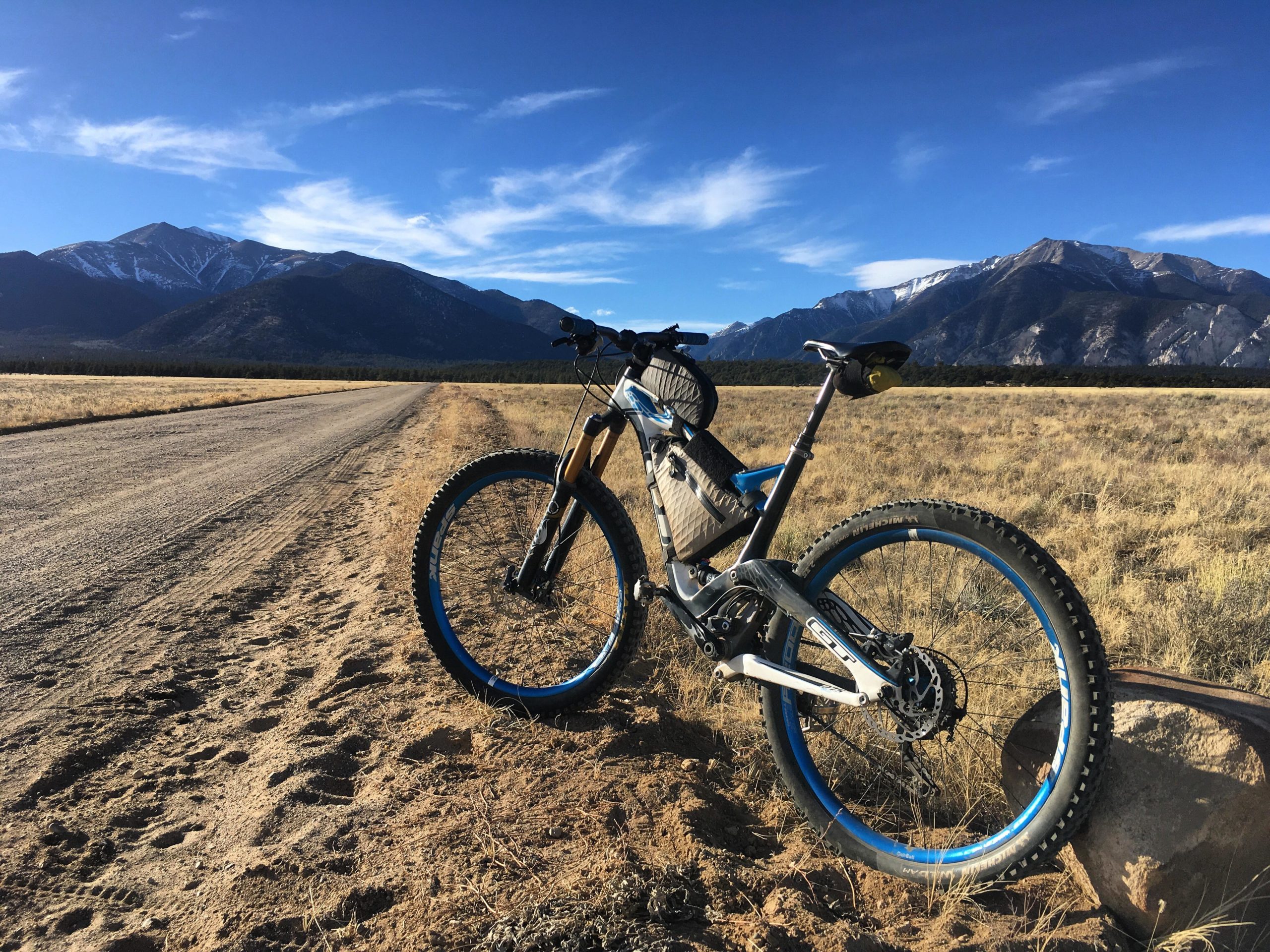 A mountain bike parked along a dirt road, with a backdrop of snow-capped mountains and a clear blue sky. The foreground features sandy terrain and sparse vegetation. The scene captures a peaceful outdoor setting ideal for biking and exploring nature. Browns Creek Road / #272 mountain bike trail.