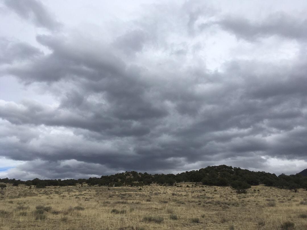 A wide-open landscape featuring a dry, grassy plain with scattered shrub vegetation, under a dramatic sky filled with dark, swirling clouds. Midland Hills Trails mountain bike trail.