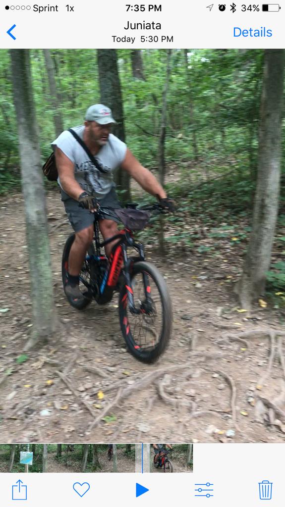 Giant FULL-E  0 SX PRO: A person riding a mountain bike on a narrow dirt trail surrounded by trees. The individual is wearing a cap and casual clothing while navigating through a wooded area. The bike is equipped with thick tires, and the scene captures the motion of the ride.