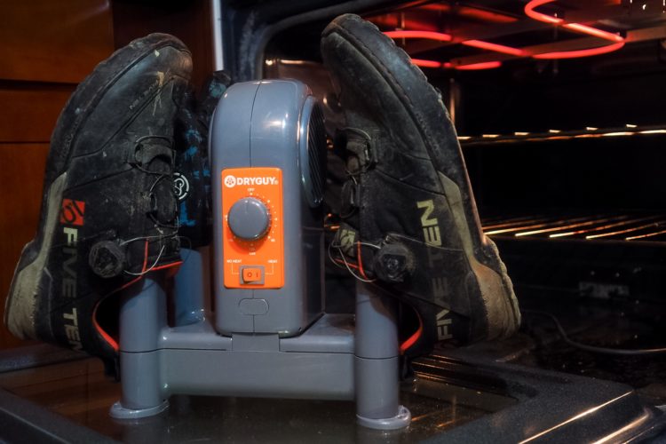 A pair of dirty hiking shoes placed on a shoe dryer, with the dryer unit's control settings visible, and an oven in the background.