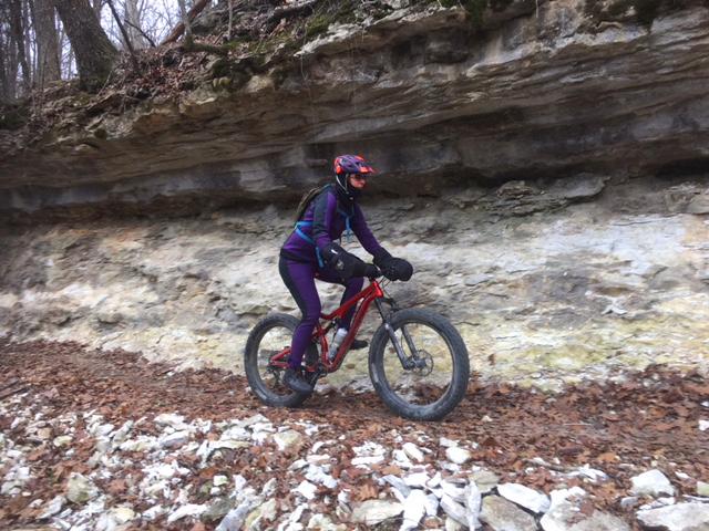 A person riding a mountain bike along a rocky trail next to a large stone cliff, surrounded by fallen leaves and trees in a wooded area. The rider is wearing a helmet and colorful cycling attire. Slaughter Pen Trail mountain bike trail.