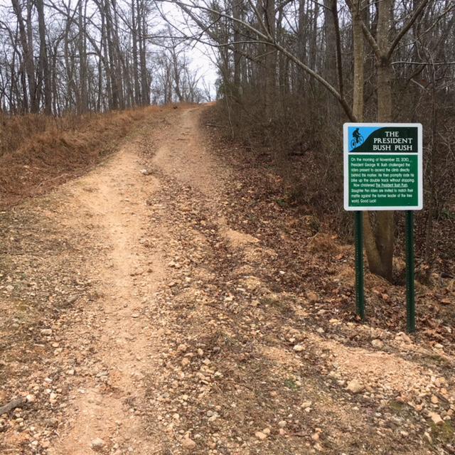 A dirt path leading uphill, flanked by bare trees and brush, with a sign on the right side labeled "The President Bush Push." The sign provides information about the trail and its history. The scene is set in a natural, wooded environment during a cloudy day. Slaughter Pen Trail mountain bike trail.