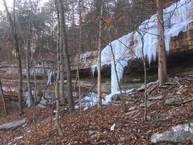 A rocky hillside is partially covered with icicles hanging from a cliff, surrounded by leafless trees and fallen leaves on the forest floor. The scene captures the beauty of winter in a wooded area. Blowing Springs mountain bike trail.