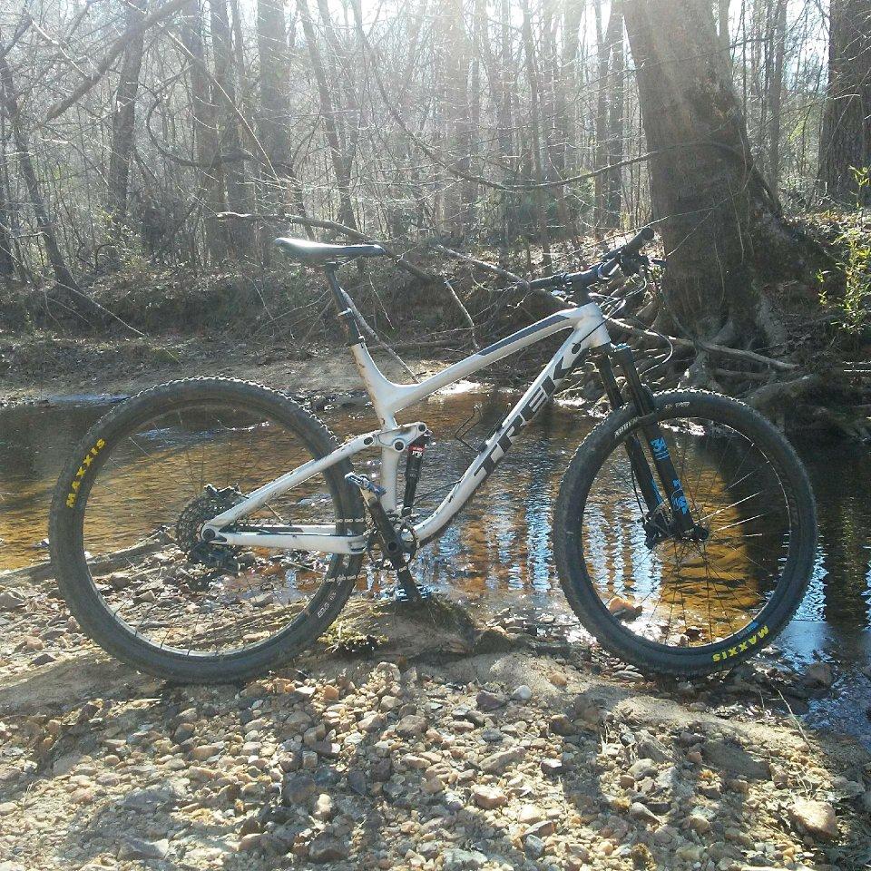 Trek Fuel EX 9 29: A mountain bike in a natural setting near a small stream, surrounded by trees and dappled sunlight. The bike is predominantly white with black and blue details, positioned on a rocky bank with water reflections visible in the background.