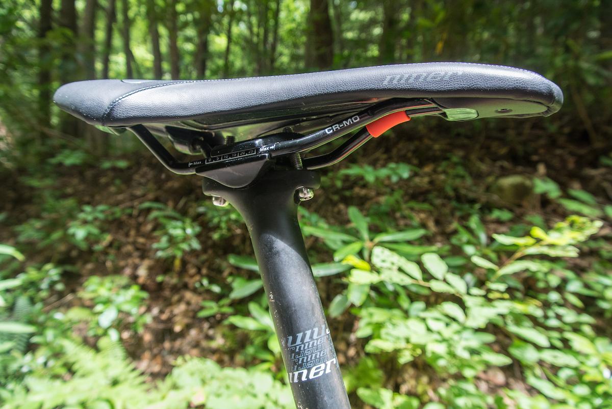 Niner Air 9 RDO: Close-up view of a bicycle saddle and seat post, set against a blurred background of green foliage and trees. The saddle features a sleek design with markings for adjustment, while the seat post is black with branding visible.