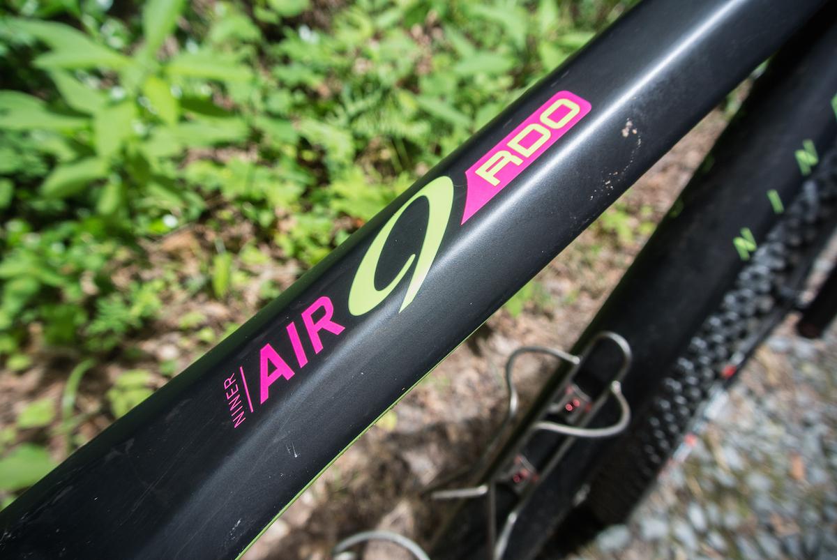 Niner Air 9 RDO: Close-up of a bicycle frame with the branding "Niner AIR 9 RDO" displayed prominently. The frame is black with green and pink accents, set against a blurred background of greenery and gravel.