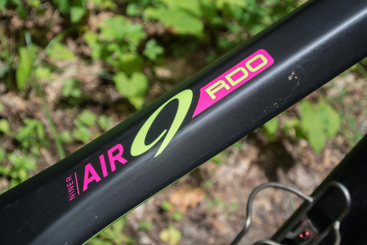 Niner Air 9 RDO: Close-up view of a black bicycle frame featuring a distinctive logo that reads "Niner AIR 9 RDO" in vibrant pink and green colors, set against a blurred background of greenery.
