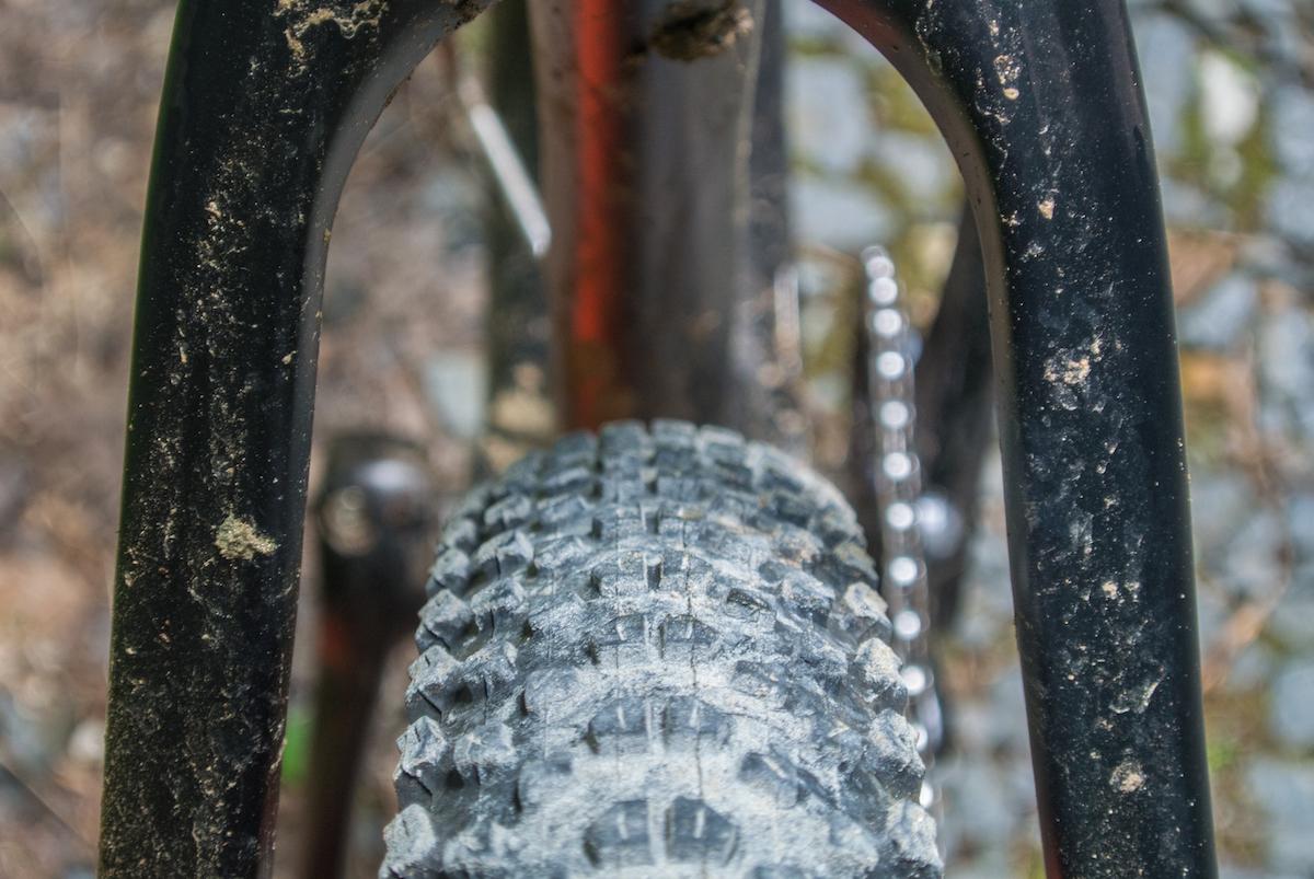Niner Air 9 RDO: A close-up view of a mountain bike's rear wheel and frame, featuring a heavily treaded tire, dirt and mud deposits on the bike's components, and a blurred background of gravel and foliage.