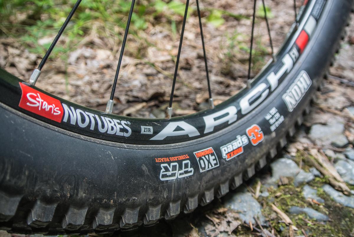 Niner Air 9 RDO: Close-up of a mountain bike tire and rim resting on a forest floor. The tire shows a tread pattern, and the rim features branding from Stan's NoTubes and various stickers, including "ZTR" and "Maxxis." The background consists of leaves and gravel, emphasizing an outdoor setting.
