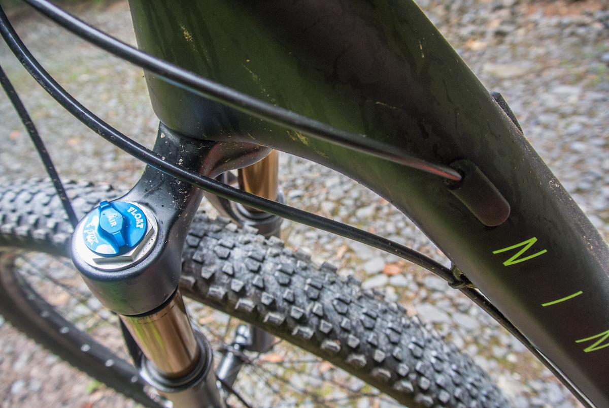 Niner Air 9 RDO: Close-up view of a mountain bike's front suspension system, featuring a blue adjustment knob labeled "FLOAT." The bike frame is black with green accents, and the textured tire tread is visible in the background, set against a gravel surface.