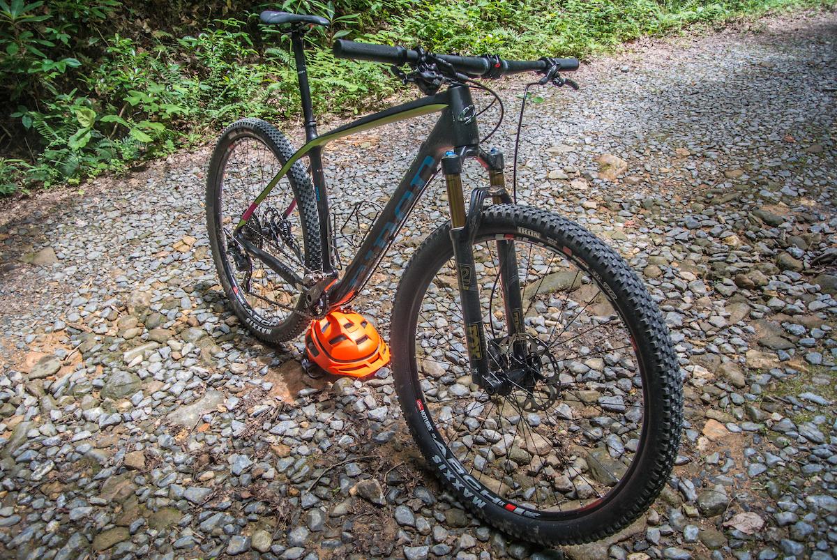 Niner Air 9 RDO: A mountain bike with a black frame and vibrant accents is parked on a gravel path surrounded by greenery. An orange helmet rests on the ground beside the bike, suggesting it is ready for a ride on the trail.