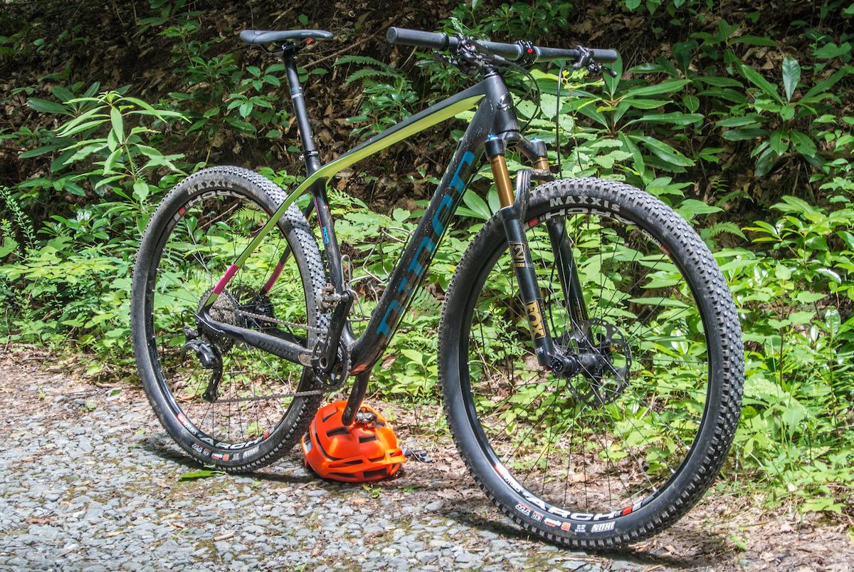 Niner Air 9 RDO: A mountain bike with a lightweight frame and thick, treaded tires is parked on a gravel trail surrounded by green foliage. An orange helmet sits at the base of the bike. The bike features high-end components visible on the frame and wheels, suggesting it's designed for off-road cycling.