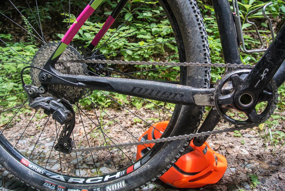 Niner Air 9 RDO: Close-up of a mountain bike's rear drivetrain, featuring a black Niner crankset, derailleur, and a cassette, with a visible chain resting on the gears. An orange helmet is placed on rocky ground in the background, surrounded by lush greenery.