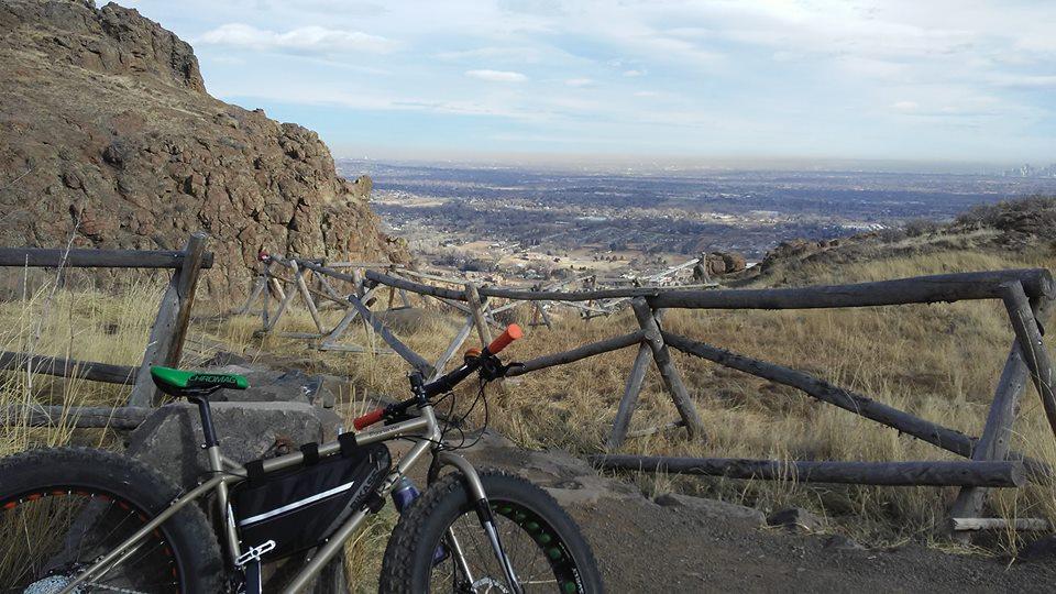 A scenic view of a hilly landscape with a foreground featuring a mountain bike resting near a wooden fence. The background showcases a vast valley extending towards the horizon, dotted with houses and under a partly cloudy sky. The terrain is dry with patches of grass and rocky formations. North Table Mountain mountain bike trail.