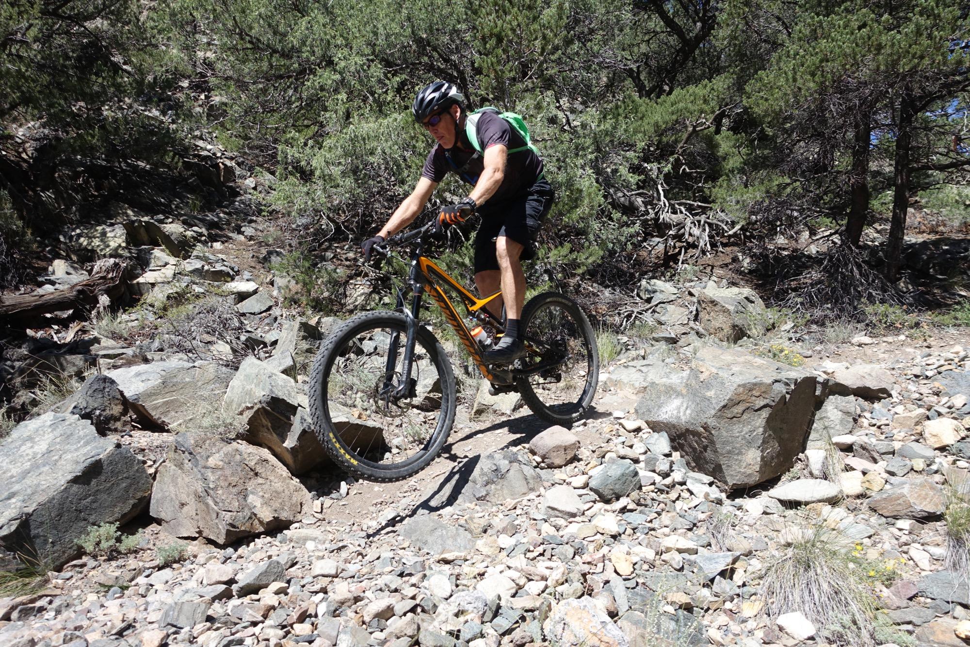 A mountain biker navigating a rocky trail, skillfully maneuvering over large stones and loose gravel, surrounded by dense greenery and trees in a natural outdoor setting. Cottonwood mountain bike trail.