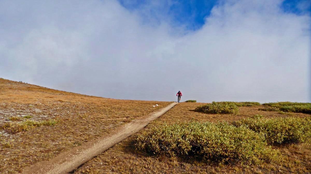 A person walking along a dirt path through a grassy, elevated landscape, with patches of low shrubs and a cloudy blue sky in the background. Monarch Crest Trail mountain bike trail.