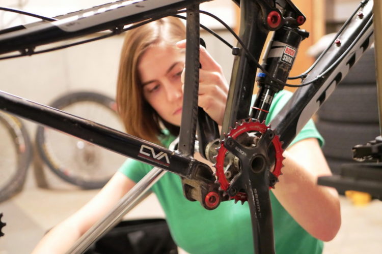 A person working on a bicycle frame in a garage setting, focusing on the drivetrain components. Various bike wheels are visible in the background. The individual is wearing a green shirt and appears concentrated on the task.