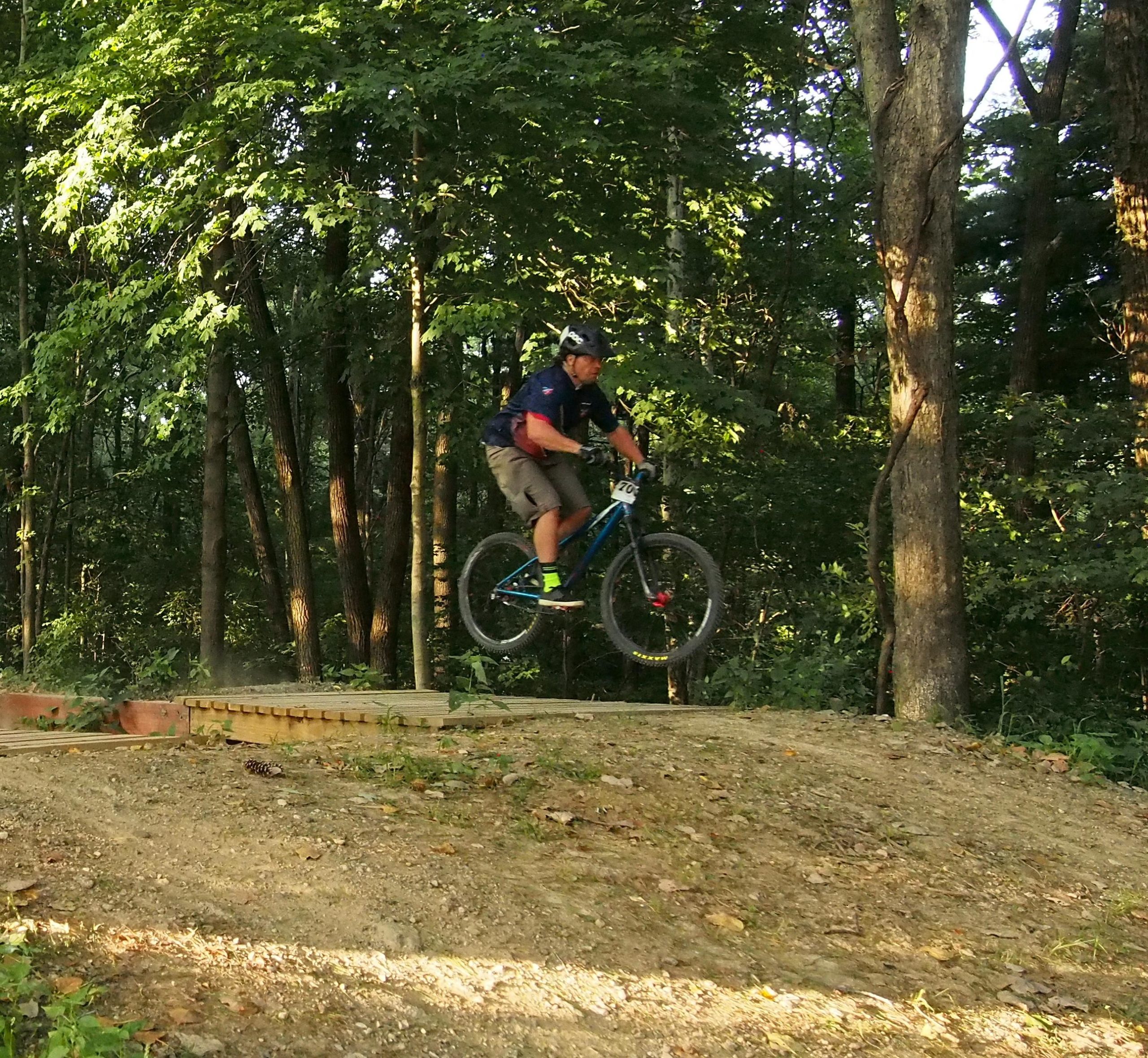 A mountain biker in action, jumping off a wooden ramp in a forested area. The biker is wearing a helmet and protective gear, with a vibrant blue bike. Sunlight filters through the trees, casting dappled shadows on the ground. Griffin Bike Park mountain bike trail.