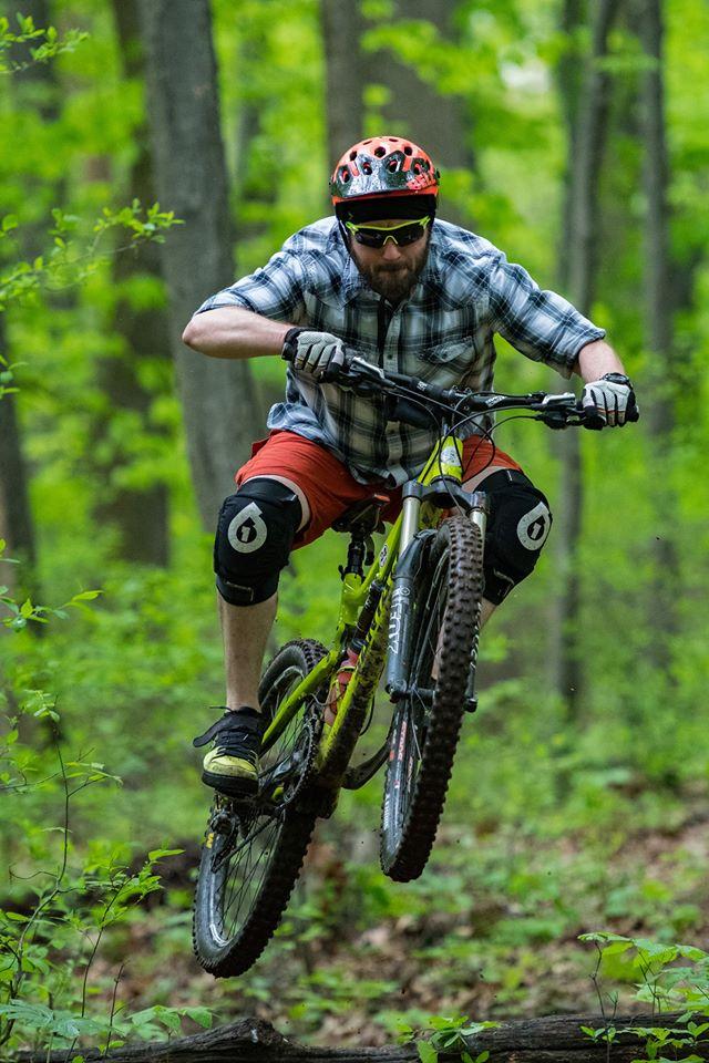 Santa Cruz Bronson: A mountain biker wearing a red helmet and sunglasses performs a jump over a log in a wooded area. He is dressed in a plaid shirt and orange shorts, with knee pads for protection, surrounded by lush green trees and foliage.
