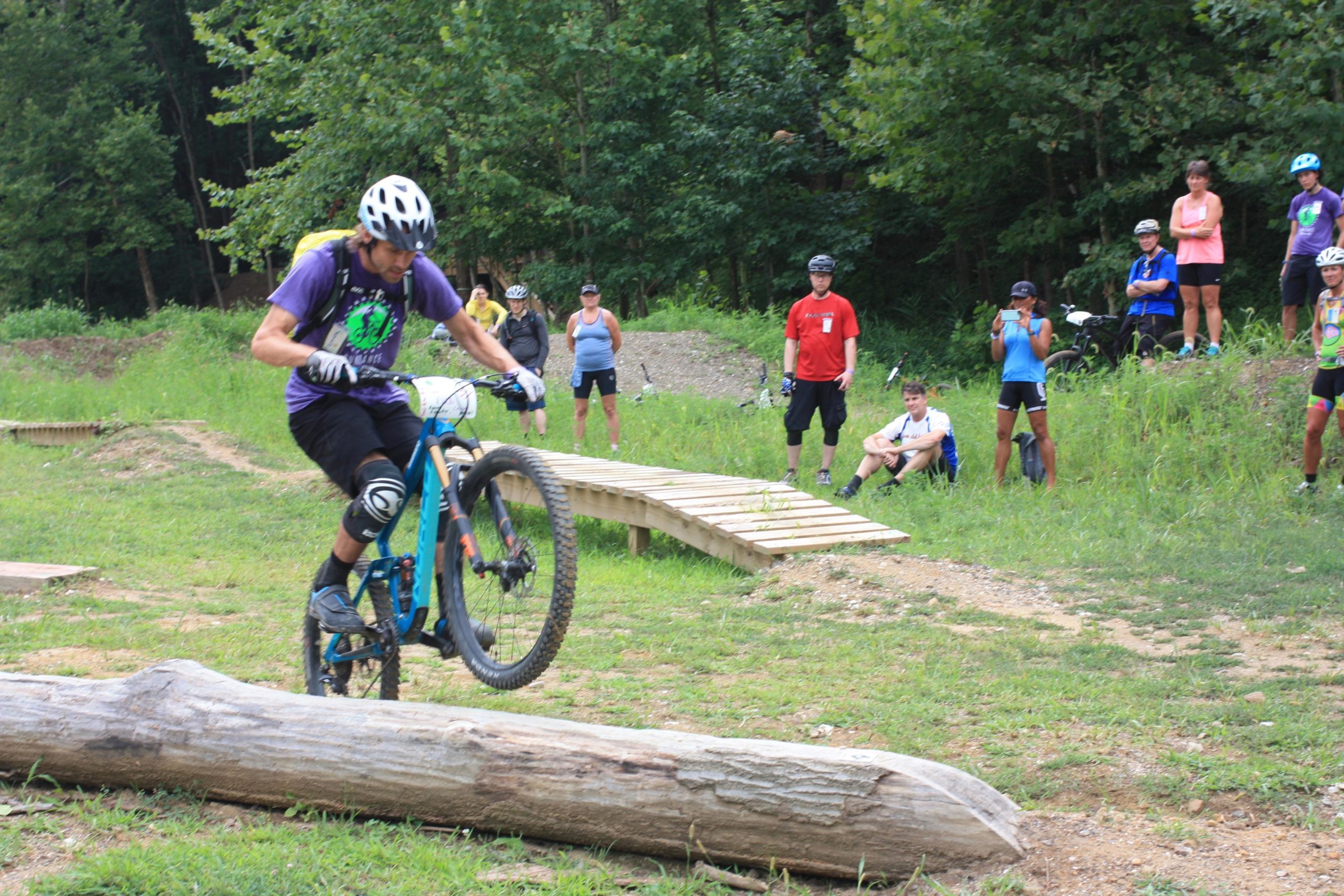 A mountain biker in a purple shirt and helmet is performing a jump over a log in a grassy area, while a group of spectators watches from the side. Some onlookers are taking photos, and a wooden ramp is visible in the background. The setting is surrounded by trees, indicating an outdoor biking event or practice session. Griffin Bike Park mountain bike trail.