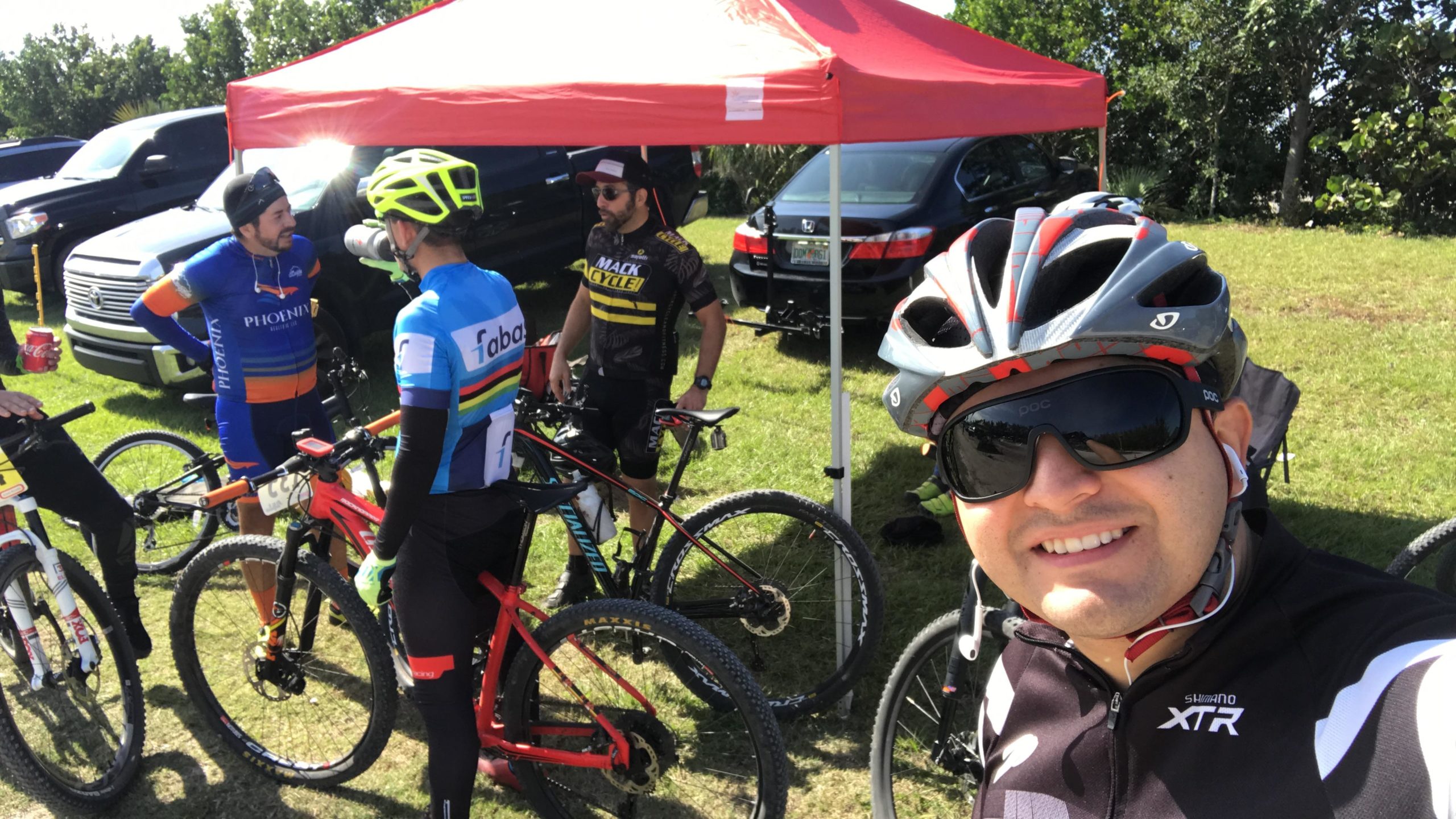 A group of four cyclists, wearing colorful cycling jerseys and helmets, gather near a red tent in a grassy area. One cyclist takes a selfie, showcasing a friendly atmosphere among the riders. Bicycles are leaned against each other, with vehicles visible in the background under bright, sunny weather. Virginia Key North Point mountain bike trail.