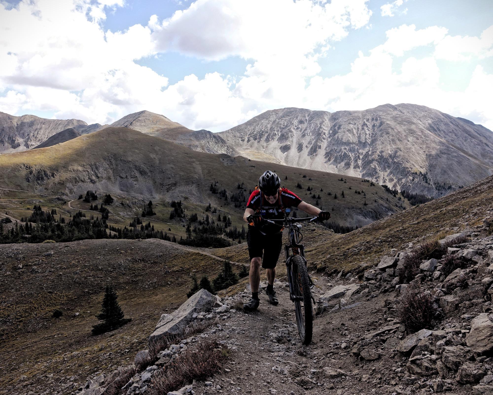A mountain biker pushing their bike up a rocky trail, surrounded by mountains under a partly cloudy sky. The terrain is rugged, with rocky paths and sparse vegetation, showcasing a dramatic alpine landscape. Canyon Creek Trail mountain bike trail.