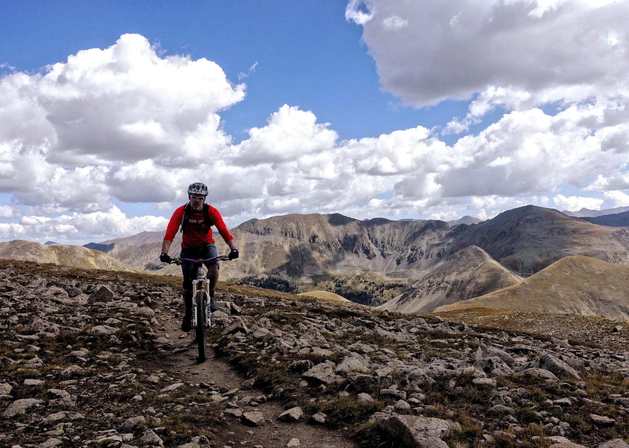 A mountain biker rides along a rocky trail with a scenic backdrop of mountains and a blue sky dotted with fluffy white clouds. The cyclist is wearing a helmet and a red long-sleeve shirt, navigating the terrain amidst a rugged landscape. Canyon Creek Trail mountain bike trail.