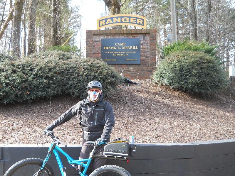 Cannondale Bad Habit: A person wearing a helmet and face covering stands next to a mountain bike at the entrance of Camp Frank D. Merrill, which features a sign reading "RANGER" above the camp name and details about the 5th Ranger Training Battalion. The background includes trees and shrubs typical of a forested area.